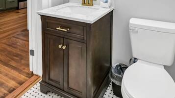 Ein Badezimmer mit einem dunklen Holz-Waschtisch, einem weißen Waschbecken, goldenen Armaturen und einem großen Spiegel mit goldener Rahmung. Es gibt eine weiße Toilette und einen kleinen Mülleimer.