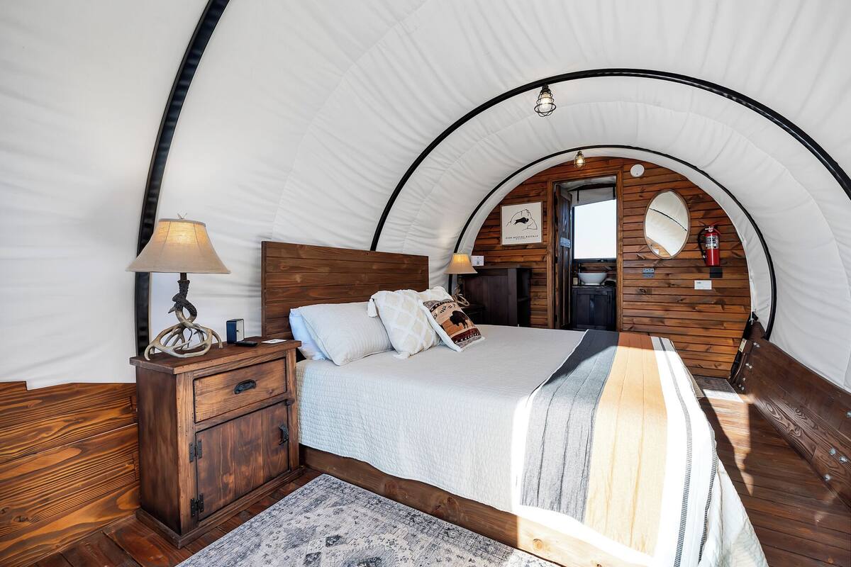 A cozy, arched tent-style bedroom with a wooden headboard, a bedside table with a lamp, and a bed with a striped blanket.