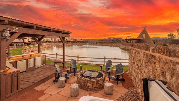 A patio area with Adirondack chairs, a stone fire pit, and a view of a lake at sunset. - Zion White Bison (Virgin)