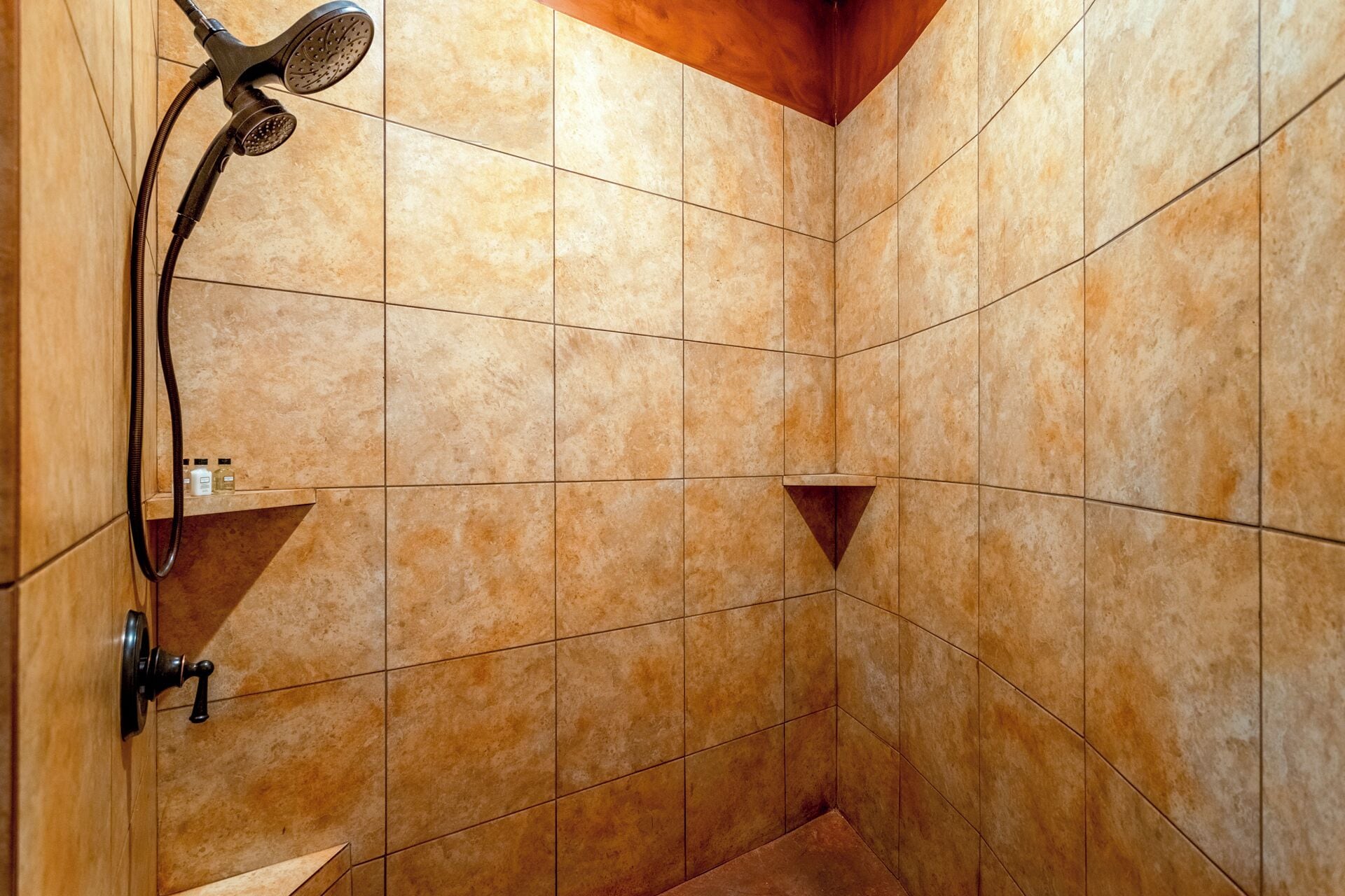 A shower area with beige tiles, a handheld showerhead, and a small shelf with toiletries.