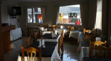 Un restaurante con varias mesas y sillas, un televisor montado en la pared y ventanas amplias con vista a la calle.