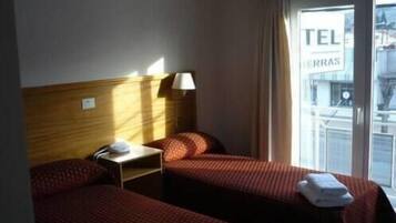 Habitación de hotel con dos camas, una mesita de noche, una lámpara y vistas al exterior por una ventana.