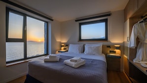 Une chambre d’hôtel avec un grand lit, deux tables de chevet avec des lampes, une vue sur la mer au coucher du soleil et une armoire en bois.