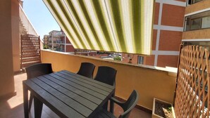 Un balcon avec une table et des chaises en bois, un auvent à rayures et une vue sur les bâtiments en arrière-plan.