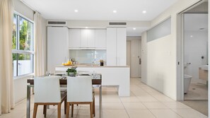 A modern kitchen with white cabinetry, a dining area with a table and chairs, and a view of the outdoors through a large window.
