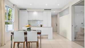 Une cuisine moderne avec des meubles blancs, un coin repas comprenant une table et des chaises, et une vue sur l’extérieur grâce à une grande fenêtre.
