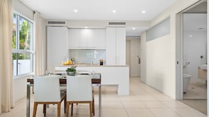 A modern kitchen with white cabinetry, a dining area with a table and chairs, and a view of the outdoors through a large window.