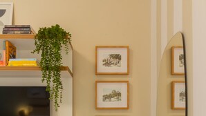Un dormitorio con cama, televisión, estante con libros y una planta, cuadros en la pared y un espejo.