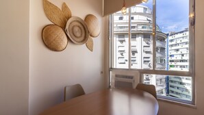 Un comedor moderno con una gran mesa de madera, vistas a un paisaje urbano a través de un ventanal y decoraciones tejidas en la pared.