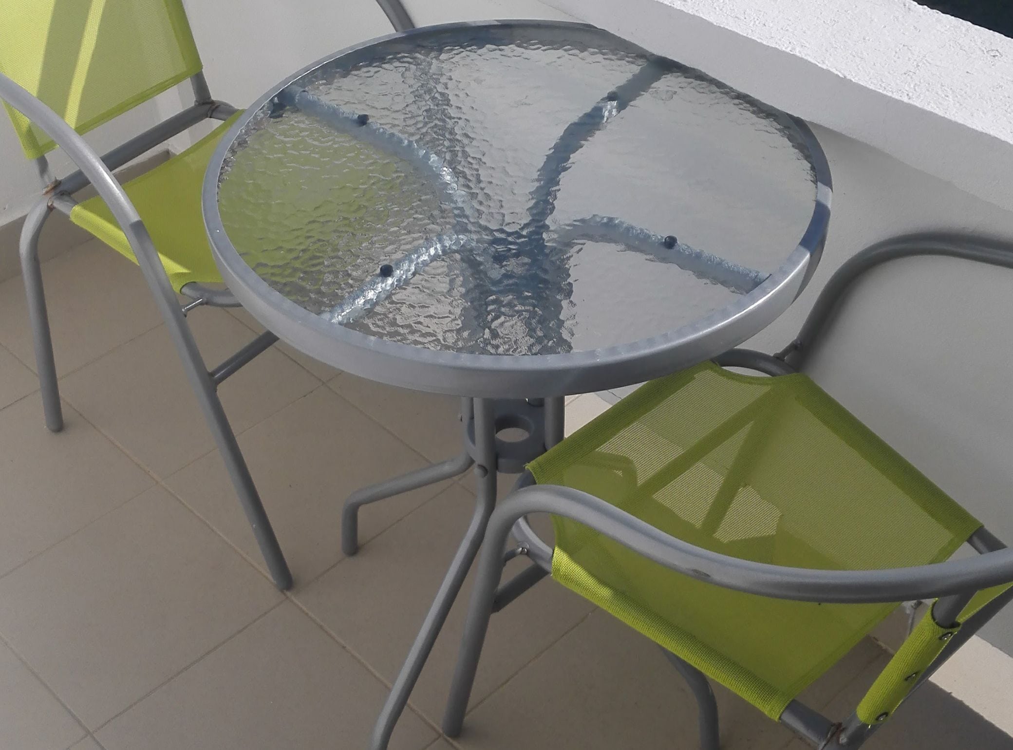 Mesa de vidro redonda com estrutura metálica, acompanhada por duas cadeiras de tela verde.