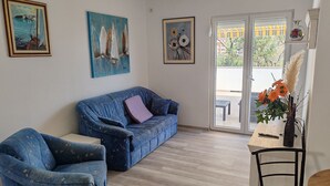 A living room with a blue sofa, a matching armchair, and a wooden coffee table. There are three paintings on the wall, a vase with flowers, and a view of the outside through a sliding glass door. - Villa Mery (Rab)