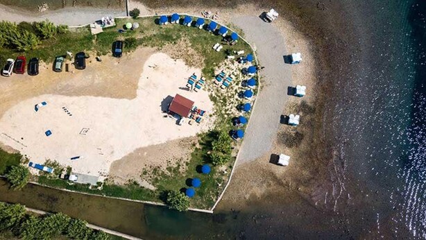 An aerial view of a beachside property with a heart-shaped layout, featuring a central building, multiple beach umbrellas, and a row of parked cars along the shoreline. - Villa Mery (Rab)