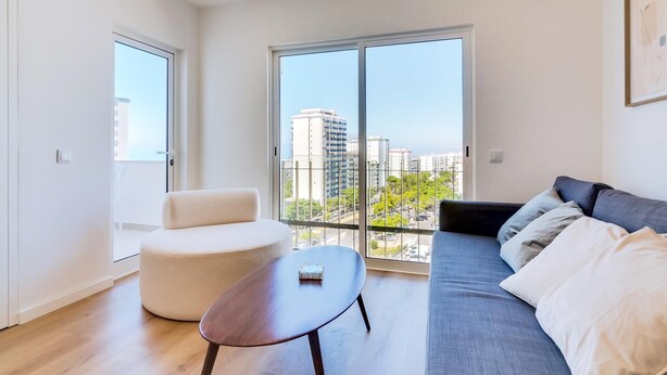 Un moderno salón con un sofá, un reposapiés redondo y una mesita. Ventanas amplias ofrecen vistas del paisaje urbano.