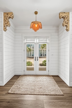 A room with white paneled walls, a wooden floor, a woven rug, and a door with a view to a blue house. - The Oyster Cottage 580 SOU (Port Aransas)
