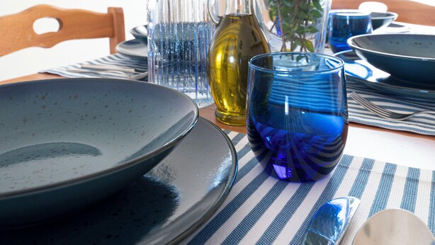 Una mesa de comedor con platos, cubiertos, una botella de vidrio, un florero azul con flores y un mantel a rayas.