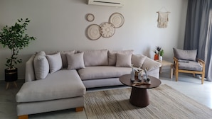Ein modernes Wohnzimmer mit einem beigefarbenen Ecksofa, einem runden Couchtisch und einem Holzstuhl. An den Wänden befinden sich dekorative Wandbehänge und ein Topf mit einer Pflanze.