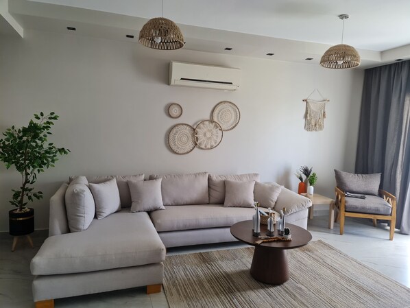 Een moderne woonkamer met een beige modulaire bank, een ronde salontafel en een houten stoel. Er zijn decoratieve wanddecoraties en een potplant.