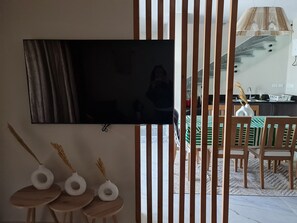 Un salon moderne doté d’un grand téléviseur à écran plat fixé au mur, d’une table à manger en bois avec des chaises, et d’un coin cuisine visible à travers une cloison.