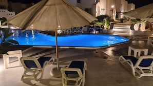Ein Poolbereich mit Liegestühlen und Sonnenschirmen, umgeben von weißen Gebäuden mit Rundbogenfenstern.