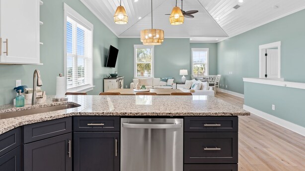 A modern kitchen with dark cabinets, a granite countertop, and a dishwasher. There is a dining area with a sofa and a TV mounted on the wall. - Seaside Escape  Walk to Beach  Private Pool + Hot Tub (Panama City Beach)