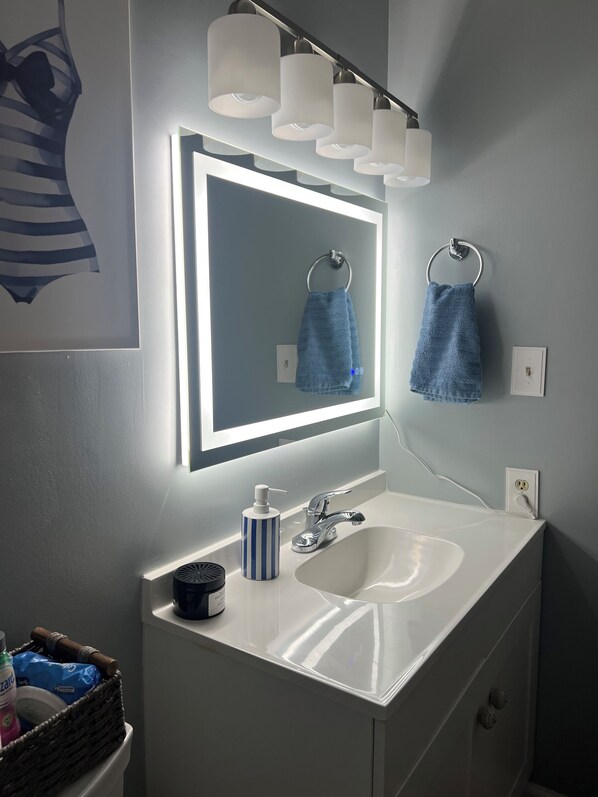 Un baño moderno con un lavamanos blanco, un espejo con iluminación integrada y dos toallas colgadas en aros.