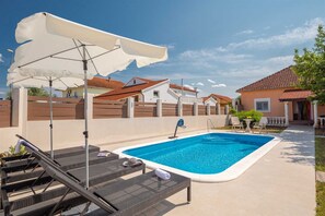 A swimming pool with lounge chairs and umbrellas, surrounded by houses with red-tiled roofs.
