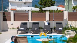 A poolside area with lounge chairs and umbrellas, a clear blue pool, and a view of buildings and trees in the background. - Villa Josefina – Heated Pool Holiday Home near Split, Trilj (Košute)