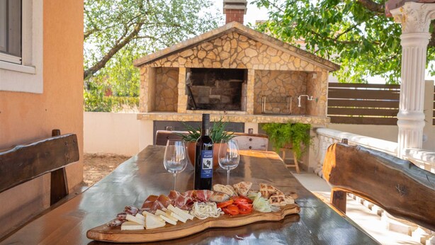 An outdoor dining area with a stone fireplace, a wooden table with a cheese platter, and a bottle of wine. - Villa Josefina – Heated Pool Holiday Home near Split, Trilj (Košute)