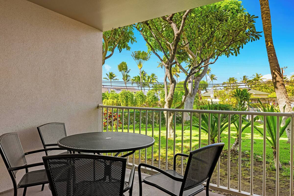 Ein Balkon mit rundem Tisch und vier Stühlen, mit Blick auf einen gepflegten Garten mit verschiedenen Bäumen und wolkenlosem, blauem Himmel.