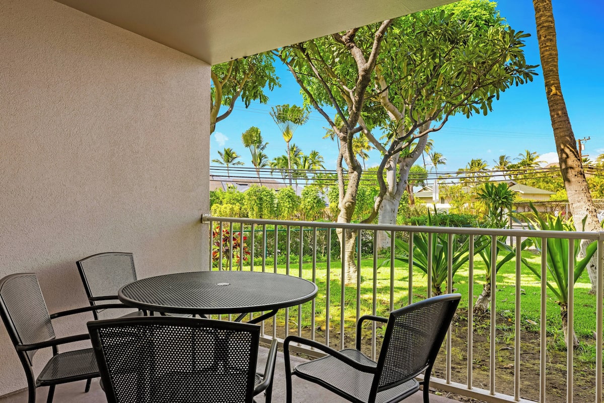 A balcony with a round table and four chairs, overlooking a well-maintained garden with various trees and a clear blue sky.