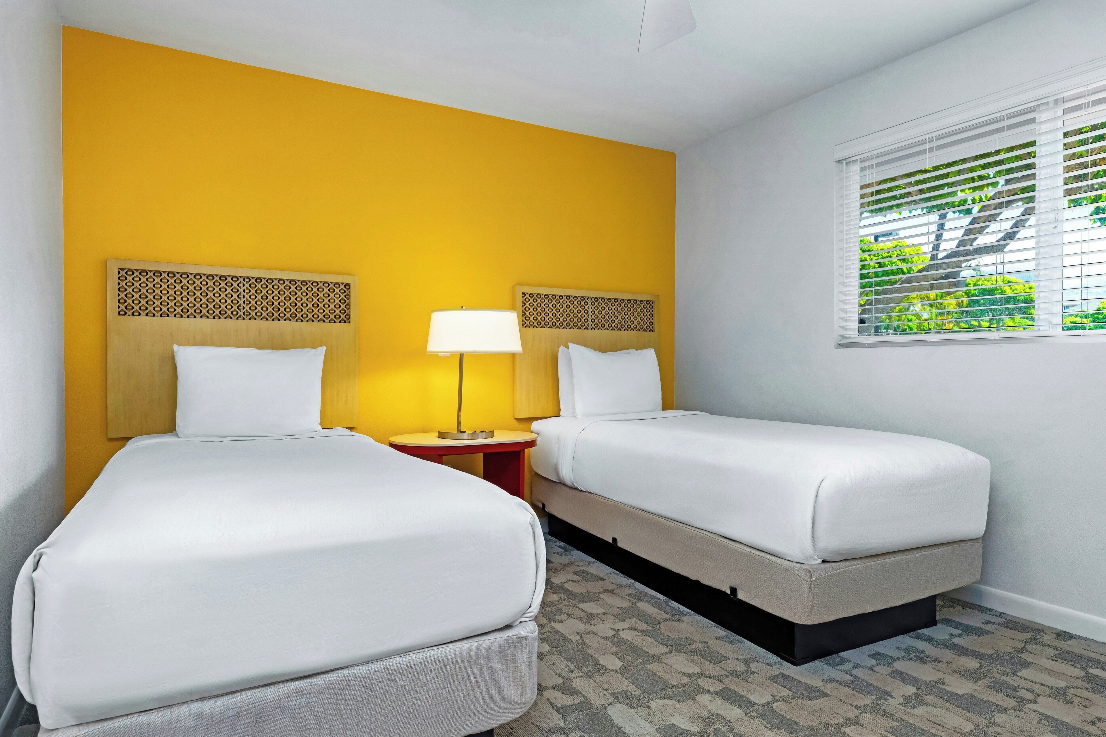 Habitación de hotel con dos camas, una mesita de noche con lámpara, una ventana con persianas y una alfombra estampada.