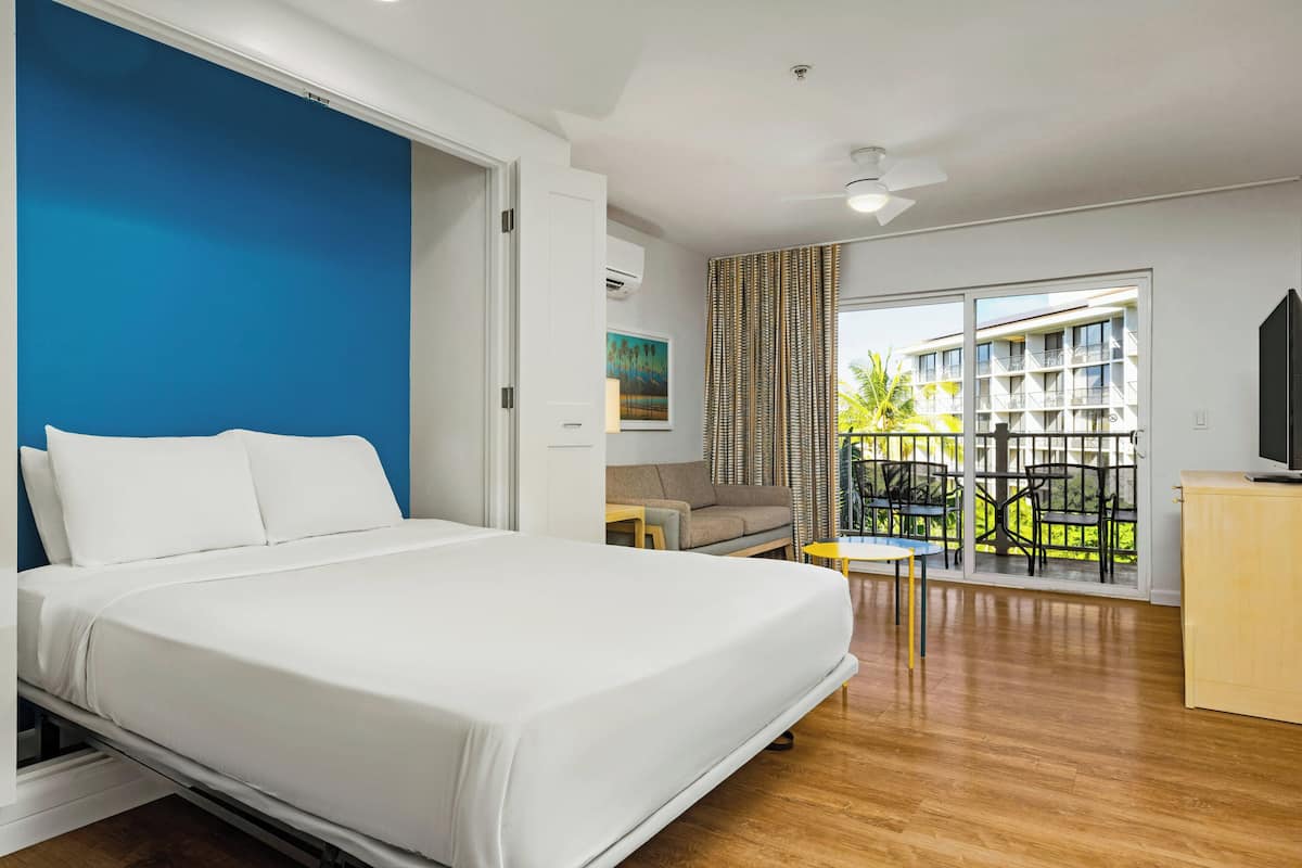 Ein Hotelzimmer mit Bett, einer Couch, einem kleinen Tisch, einem Fernseher und einem Balkon mit Blick auf Gebäude und Grünflächen.