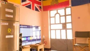Habitació amb nevera, taula de fusta, finestra amb reixes i banderes a la paret.