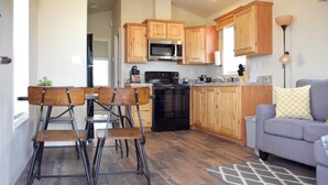 A modern kitchen with wooden cabinets, a built-in microwave, and a stove. There is a dining area with a table and chairs, and a gray sofa with patterned cushions.