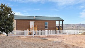 A single-story wooden cabin with a white picket fence, a covered porch, and a view of the desert landscape.