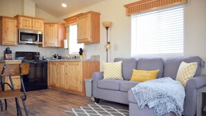 A modern kitchen with wooden cabinets, a microwave, and a stove. There is a gray sofa with yellow and white patterned pillows, a small table, and a floor lamp.