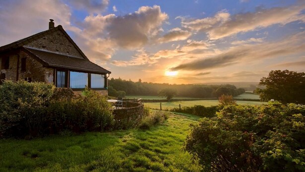 A rustic stone house with a large window, surrounded by lush greenery and a view of a sunset over a rural landscape. - Gîte du Bois de la Velle (VIRY)
