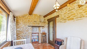 A rustic room with stone walls, a wooden ceiling, a tiled floor, a table with a floral tablecloth, a wicker chair, and a bookshelf. - Gîte du Bois de la Velle (VIRY)