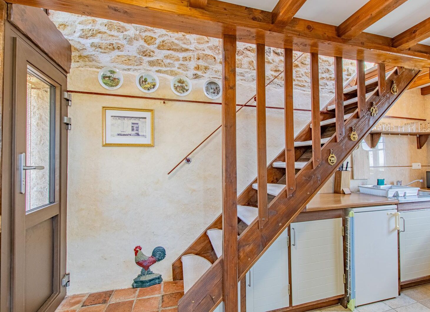 A rustic kitchen with a wooden staircase, a rooster decoration, and a stone wall with circular pictures.
