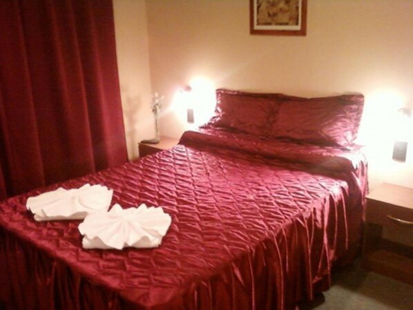 A neatly made bed with a red bedspread and pillows, a white pillow on the bed, and a nightstand with a lamp.