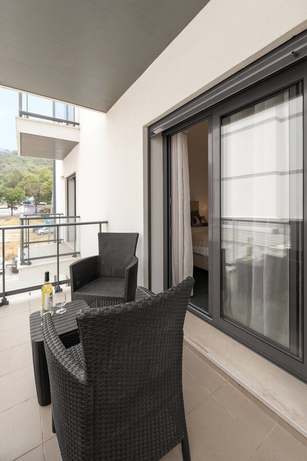 Un balcone con due sedie in vimini, un tavolo, una porta scorrevole in vetro e vista su un edificio e degli alberi.