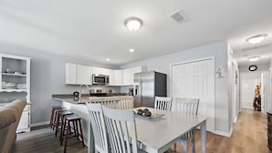 Una cocina moderna con armarios blancos, una zona de comedor con mesa y sillas de madera, y un microondas integrado.
