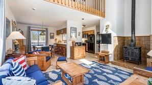 A spacious living room with a high ceiling, a wooden staircase, a kitchen area, and a black wood-burning stove. - Spacious 5-Bedroom Home Near Beach (Bethany Beach)