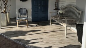 A covered porch with a blue door, a chair, a bench, a potted plant, and a wreath on the wall.