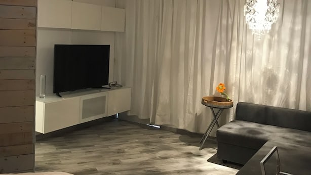 A modern hotel room with a flat-screen TV, a grey sofa, a small side table with a flower, and a large chandelier.