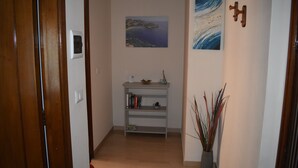 A hallway with a wooden door, a white wall, a shelf with books, a small table with a bowl, and a potted plant. - Naxos Sea Apartment (Giardini Naxos)