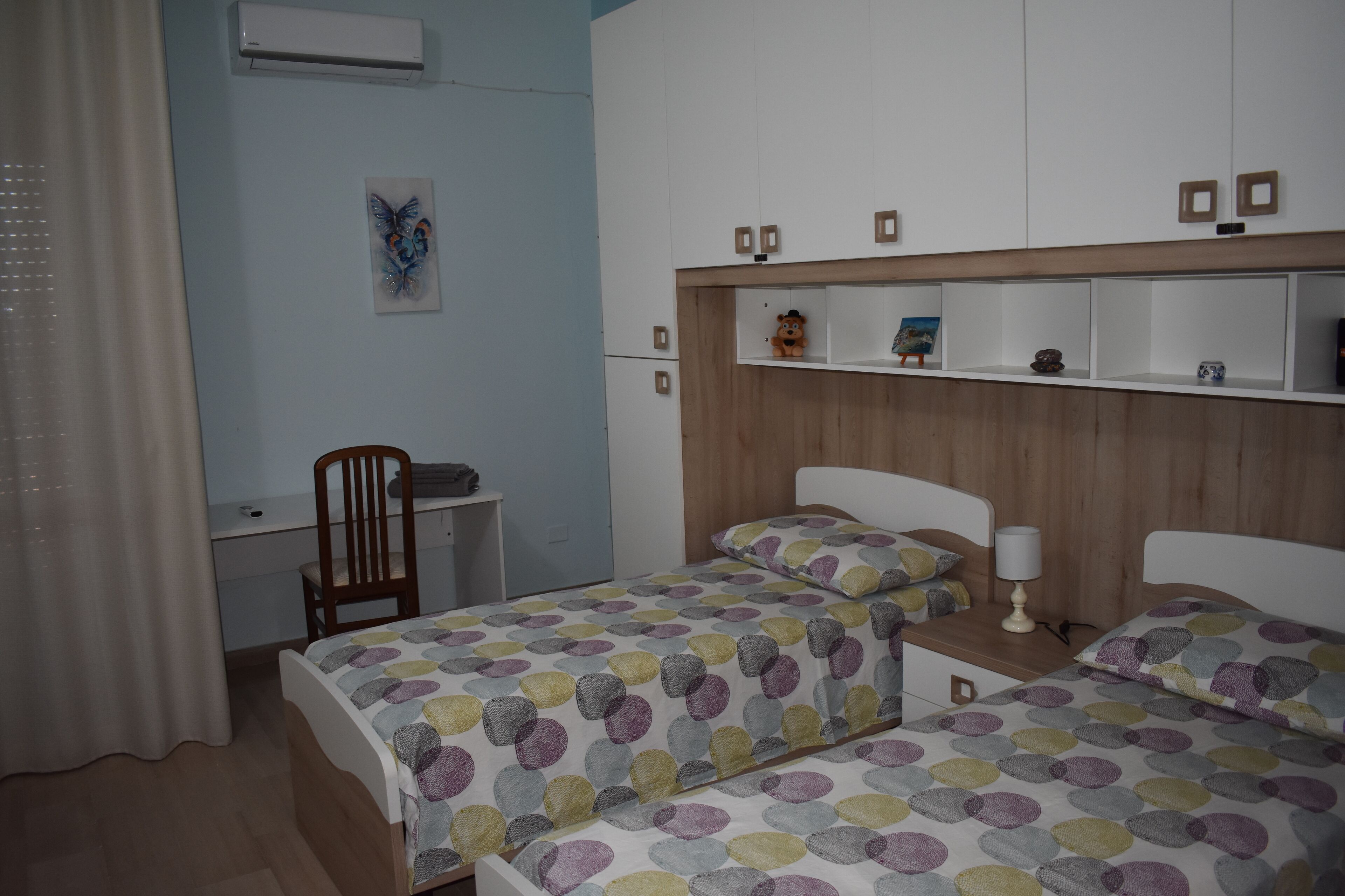 A bedroom with two beds, a desk, and a chair. There is a shelf above the beds with decorative items.
