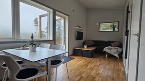 Een moderne kamer met een eettafel, stoelen, een bank, een televisie en een raam met uitzicht op bomen.