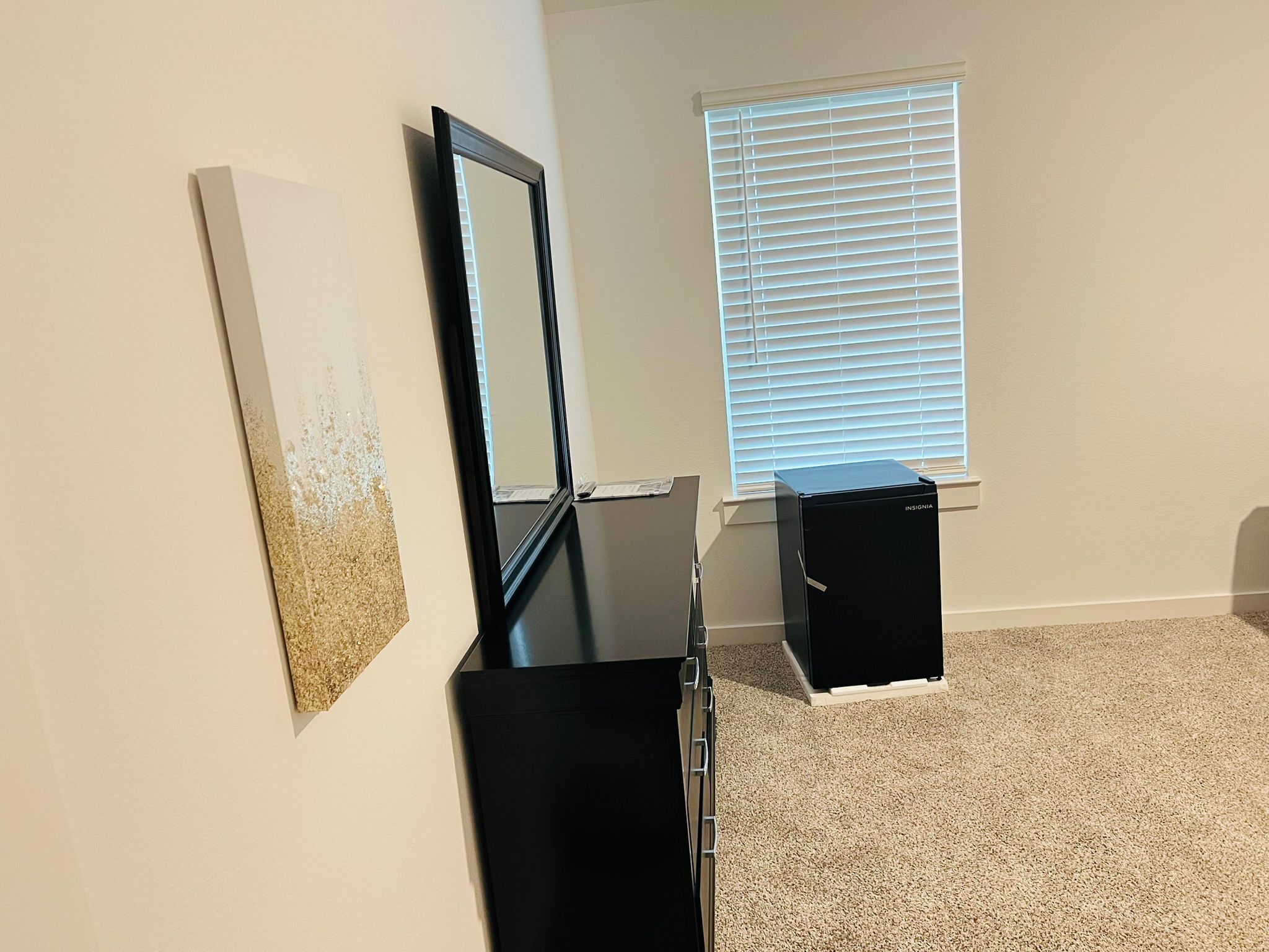 A room with a black dresser, a mirror, a framed picture on the wall, and a window with blinds.