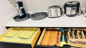 En köksbänk med en kaffebryggare, brödrost och riskokare. Under bänken finns en låda med en skärbräda, köksredskap och en stekspade.